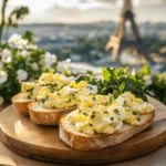 French egg salad served on toasted baguette slices with herbs, set on a wooden board with the Eiffel Tower blurred in the background.